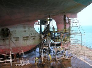Técnicos trabajando sobre andamios bajo el casco de un buque en dique, realizando trabajos de reparación y mantenimiento